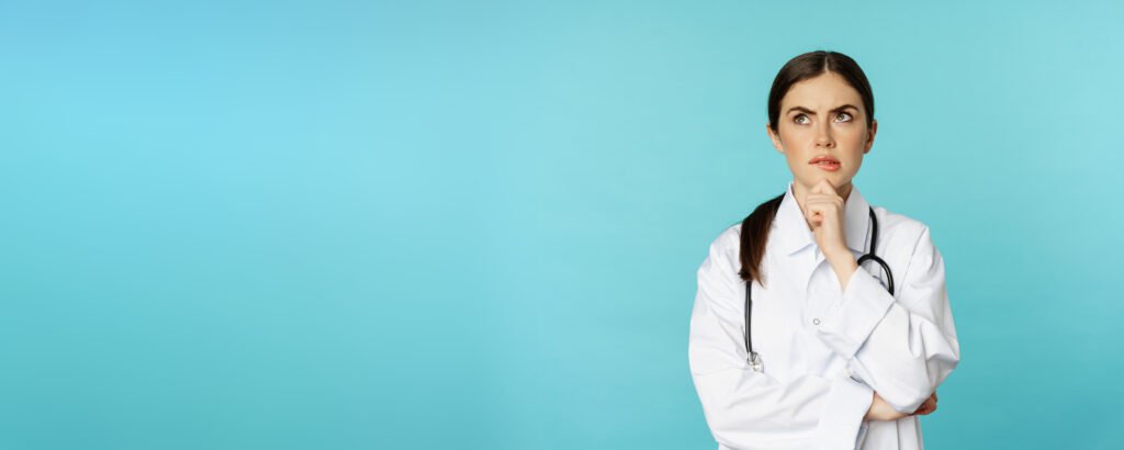 Young female doctor, hospital worker in white coat, thinking and looking away thoughtful, searching solution, standing over toquoise background